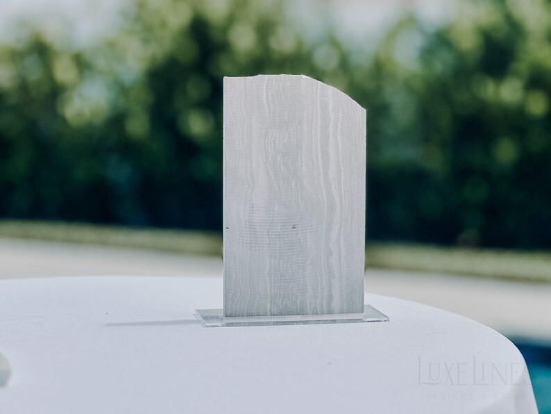 Op de afbeelding: Een rechthoekig, lichtgrijs houten tafelnummer met een gebogen bovenrand, geplaatst op een heldere acrylbasis. Het tafelnummer heeft een textuur van houtnerf. De achtergrond is wazig, met groene bladeren.