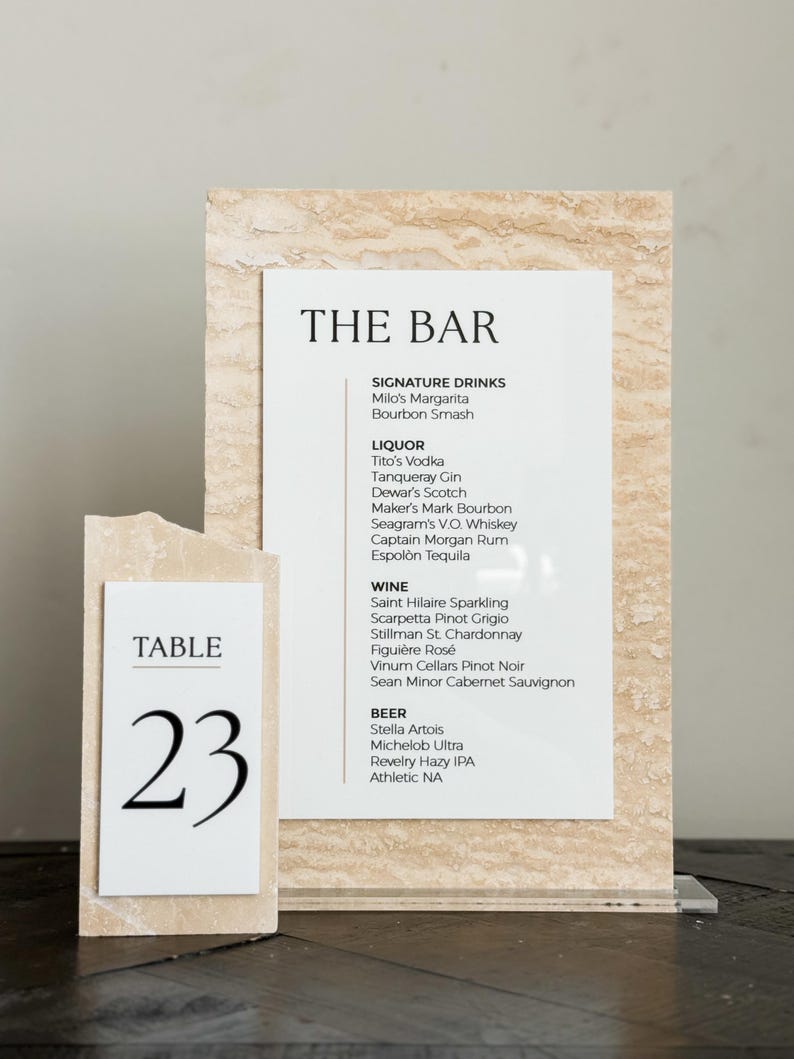 May include: A bar menu and table number 23 are displayed on a light beige stone-like background. The bar menu lists signature drinks, liquor, wine, and beer. The table number is a white card with the number 23 in a large, bold font.