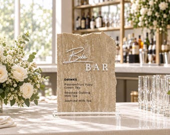 May include: A rectangular, rough-edged beige stone sign with a clear acrylic base. The sign features white text reading Boba BAR and a list of drinks: DRINKS, Passionfruit Yuzu Green Tea, Roasted Oolong Milk Tea, and Jasmine Milk Tea.