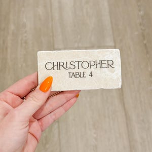 May include: A rectangular place card with the name "CHRISTOPHER" and "TABLE 4" printed in black. The card is a light beige color and has a textured surface.