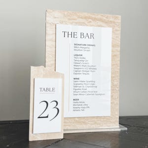 May include: A bar menu and table number 23 display. The bar menu lists signature drinks, liquor, wine, and beer. The table number is on a separate card. Both cards have a textured, light-colored background.
