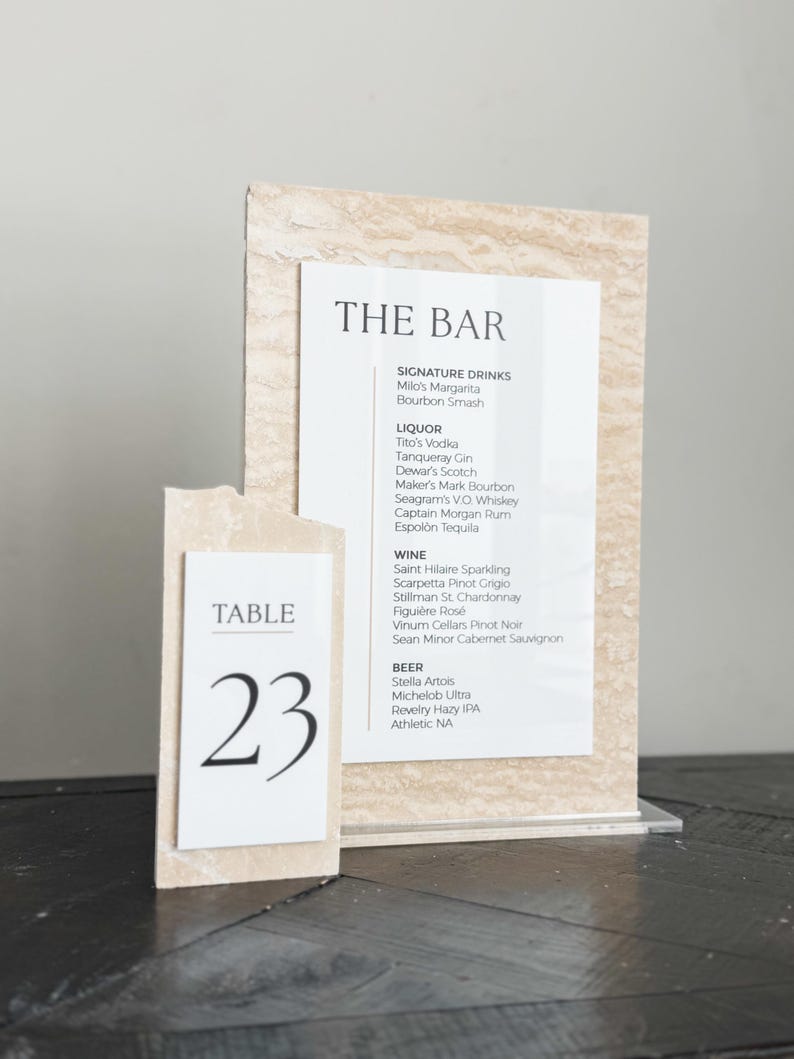 May include: A bar menu and table number 23 display. The bar menu lists signature drinks, liquor, wine, and beer. The table number is on a separate card. Both cards have a textured, light-colored background.