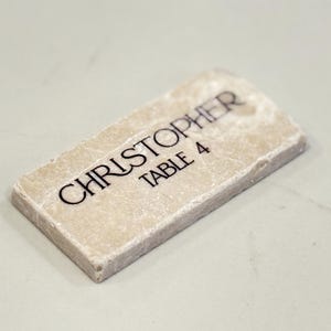 May include: A rectangular, off-white place card with the name "CHRISTOPHER" and "TABLE 4" printed in black. The place card has a textured surface and a slightly rounded top edge.