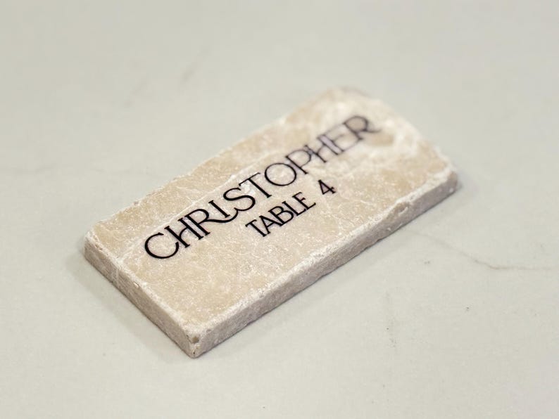 May include: A rectangular, off-white place card with the name "CHRISTOPHER" and "TABLE 4" printed in black. The place card has a textured surface and a slightly rounded top edge.