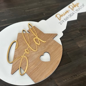 May include: A white and wood key-shaped sign with the word "Sold" in gold lettering on a house-shaped cutout. The key has a gold inscription that reads "Darian D'Amico | REALTOR | 219.869.0824".