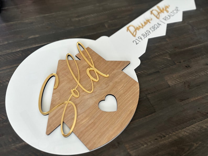 May include: A white and wood key-shaped sign with the word "Sold" in gold lettering on a house-shaped cutout. The key has a gold inscription that reads "Darian D'Amico | REALTOR | 219.869.0824".