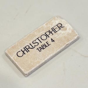 May include: A rectangular, off-white place card with the name "CHRISTOPHER" and "TABLE 4" printed in black. The card has a textured surface and a slightly rough edge.