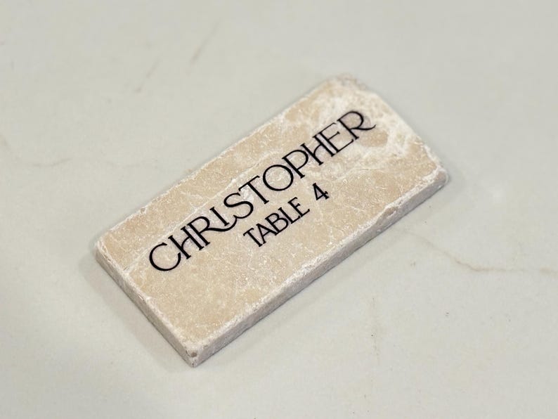 May include: A rectangular, off-white place card with the name "CHRISTOPHER" and "TABLE 4" printed in black. The card has a textured surface and a slightly rough edge.