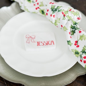 May include: A festive table setting with a white plate, a clear plate, and a napkin with a holly berry pattern. A small white card with the name "JESSICA" in red is on the plate. The napkin is knotted.