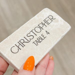 May include: A rectangular, off-white place card with the name "CHRISTOPHER" and "TABLE 4" printed in black. The card has a textured surface and is held by a hand with orange nail polish.