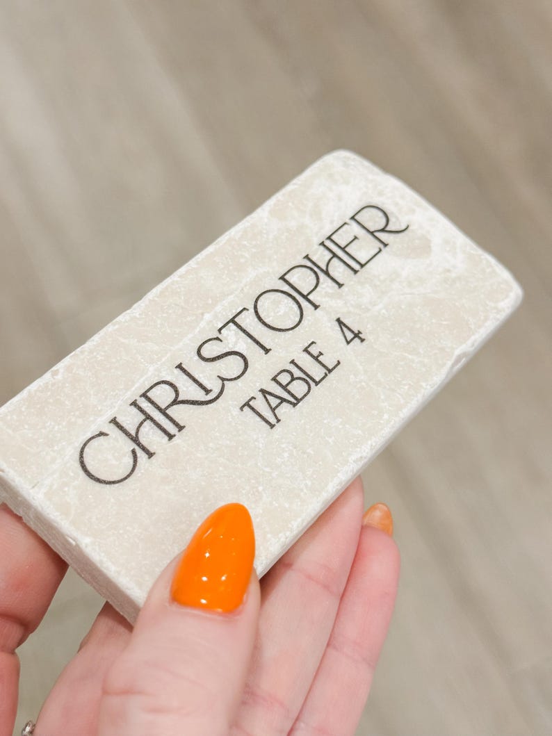 May include: A rectangular, off-white place card with the name "CHRISTOPHER" and "TABLE 4" printed in black. The card has a textured surface and is held by a hand with orange nail polish.