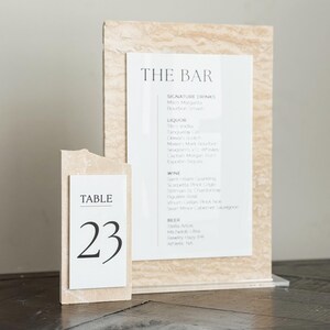 May include: A bar menu and table number 23 are displayed on a dark wooden surface. The bar menu is printed on white paper and features a list of signature drinks, liquor, wine, and beer. The table number is in a white frame.
