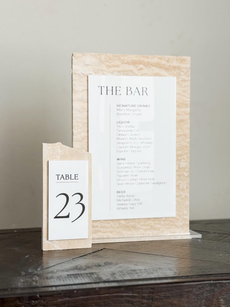 May include: A bar menu and table number 23 are displayed on a dark wooden surface. The bar menu is printed on white paper and features a list of signature drinks, liquor, wine, and beer. The table number is in a white frame.