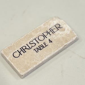 May include: A rectangular, off-white stone place card with the name "CHRISTOPHER" and "TABLE 4" in black script. The stone has a textured surface and is set against a light background.