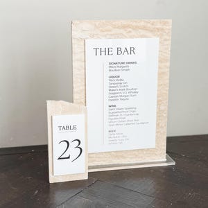 May include: A table number 23 card and a bar menu sign. The table number is a small, rectangular block with the word "TABLE" and the number "23" in black. The bar menu lists signature drinks, liquor, wine, and beer.