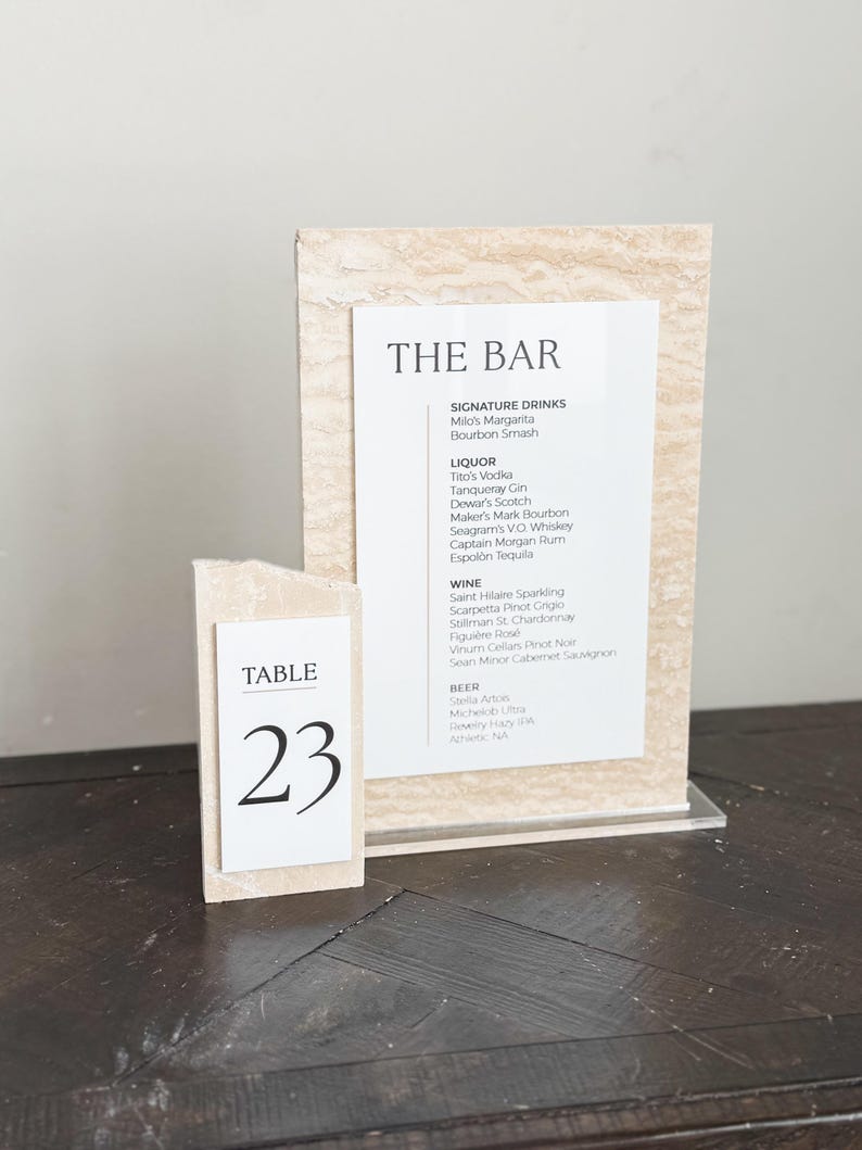 May include: A table number 23 card and a bar menu sign. The table number is a small, rectangular block with the word "TABLE" and the number "23" in black. The bar menu lists signature drinks, liquor, wine, and beer.