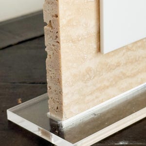 May include: A close-up of a decorative object featuring a rectangular, beige stone column with a rough, textured surface. The column is attached to a clear, rectangular acrylic base. A white panel is visible behind the stone.