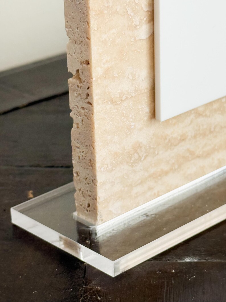 May include: A close-up of a decorative object featuring a rectangular, beige stone column with a rough, textured surface. The column is attached to a clear, rectangular acrylic base. A white panel is visible behind the stone.