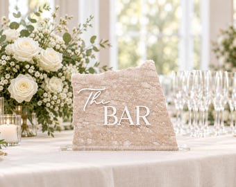May include: A beige, rough-edged stone sign featuring the white text "The BAR" in a combination of script and block lettering. The sign is mounted on a clear rectangular base and stands upright on a flat surface.