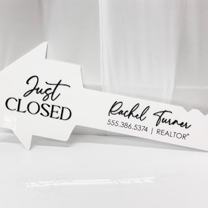 May include: A white, key-shaped sign featuring the black text "Just CLOSED" on the head and "Rachel Turner 555.386.5374 | REALTOR" on the blade. The sign is designed with a house-shaped head and a serrated edge typical of a physical key.