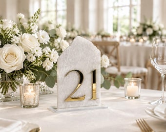 Pode incluir: Um sinal vertical de número de mesa com arco pontiagudo, feito de pedra marmoreada cinza-claro, com o numeral 21 dourado e reflexivo fixado na frente. O sinal repousa sobre uma base retangular de pedra em uma toalha de mesa branca.