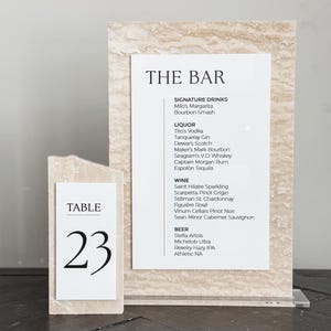 May include: A bar menu and table number 23 are displayed on a light beige stone-like surface. The bar menu lists signature drinks, liquor, wine, and beer options. The table number is in a simple black font.