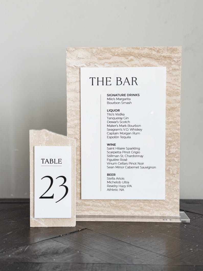 May include: A bar menu and table number 23 are displayed on a light beige stone-like surface. The bar menu lists signature drinks, liquor, wine, and beer options. The table number is in a simple black font.