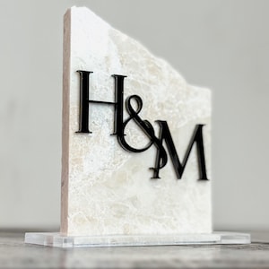 May include: A decorative marble block with the letters "H&M" in black. The marble has a natural, textured appearance and sits on a clear acrylic base. The letters are stylized and connected.