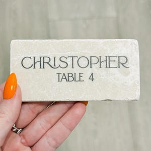 May include: A rectangular, off-white stone place card with the name "CHRISTOPHER" and "TABLE 4" printed in black. The stone has a textured surface and is held by a hand with an orange painted nail.