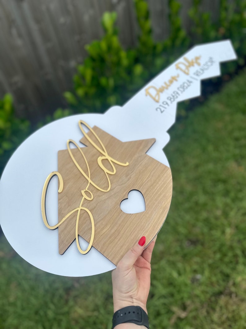 May include: A white and gold key-shaped sign with the words "Sold" and "Dawson Dhyse 219 849 1824 | Realtor" on it.