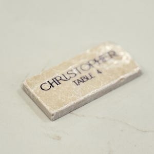 May include: A rectangular place card with the name "CHRISTOPHER" and "TABLE 4" printed in black on a light beige surface. The edges of the card have a rough, textured appearance.