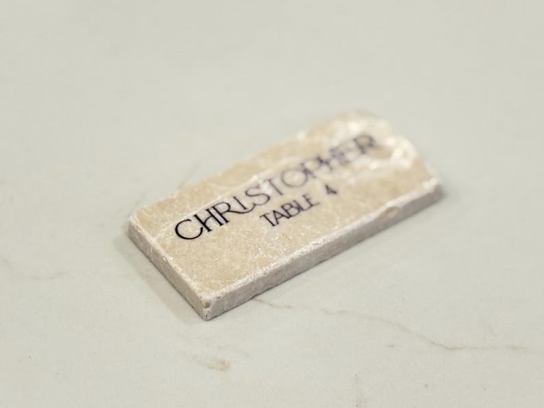May include: A rectangular place card with the name "CHRISTOPHER" and "TABLE 4" printed in black on a light beige surface. The edges of the card have a rough, textured appearance.