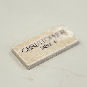 May include: A rectangular place card with the name "CHRISTOPHER" and "TABLE 4" printed in black on a beige stone-like surface. The place card has a rough, textured edge and is set against a light background.
