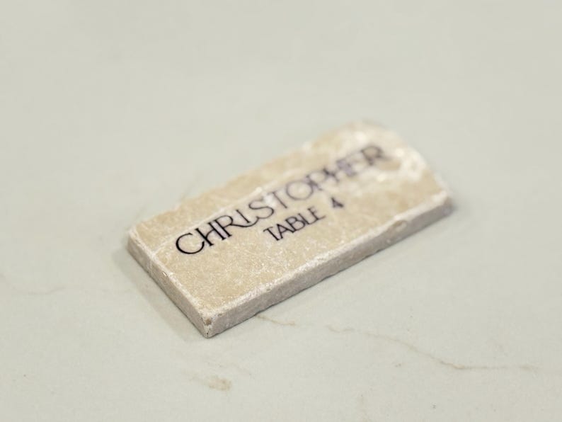 May include: A rectangular place card with the name "CHRISTOPHER" and "TABLE 4" printed in black on a beige stone-like surface. The place card has a rough, textured edge and is set against a light background.