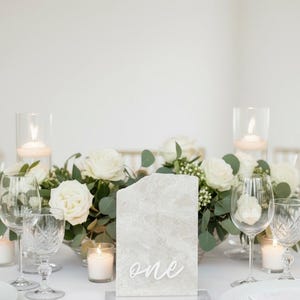 Könnte beinhalten: Elegante Tischdekoration mit weißen Rosen, Kerzen und Gläsern. Eine marmorähnliche Tischnummer mit dem Wort "one" ist zu sehen. Goldenes Besteck und weiße Teller vervollständigen die anspruchsvolle Anordnung, ideal für eine Hochzeit.