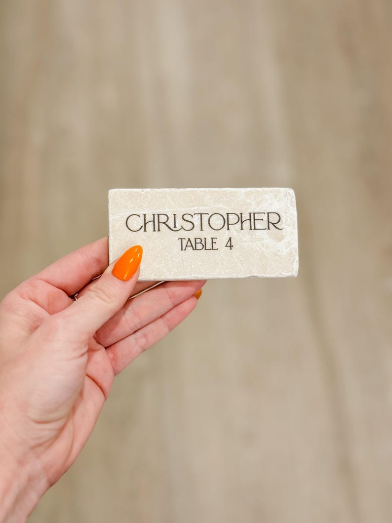 May include: A rectangular, off-white place card with the name "CHRISTOPHER" and "TABLE 4" printed in black. The card has a textured surface and is held by a hand with orange nail polish.