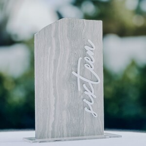 Op de afbeelding: Een rechthoekig tafelnummer met een grijze, gemarmerde uitstraling. Het woord "sixteen" staat in witte cursieve letters op de voorkant. Het tafelnummer is gemonteerd op een heldere acryl basis.