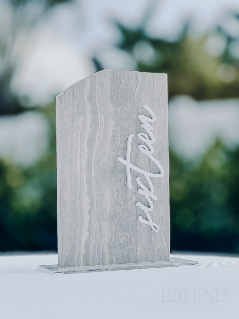 Op de afbeelding: Een rechthoekig tafelnummer met een grijze, gemarmerde uitstraling. Het woord "sixteen" staat in witte cursieve letters op de voorkant. Het tafelnummer is gemonteerd op een heldere acryl basis.