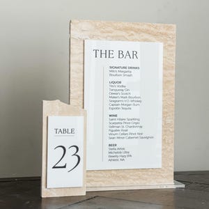May include: A bar menu and table number 23 are displayed. The bar menu lists signature drinks, liquor, wine, and beer. The table number is on a separate, smaller sign. Both signs have a beige, textured background.