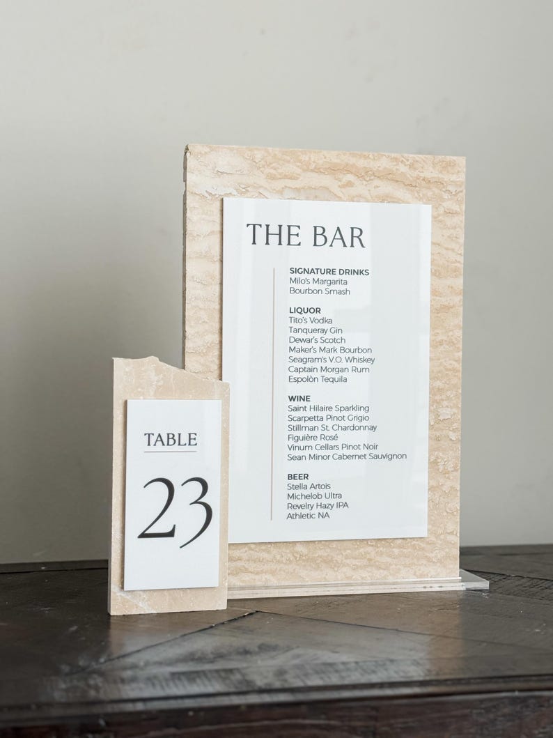 May include: A bar menu and table number 23 are displayed. The bar menu lists signature drinks, liquor, wine, and beer. The table number is on a separate, smaller sign. Both signs have a beige, textured background.
