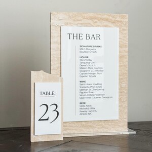 May include: A bar menu and table number 23 display. The bar menu lists signature drinks, liquor, wine, and beer. The table number is on a separate, smaller sign. Both signs have a beige, textured background.