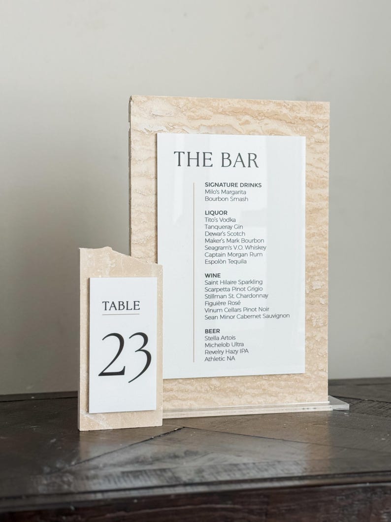 May include: A bar menu and table number 23 display. The bar menu lists signature drinks, liquor, wine, and beer. The table number is on a separate, smaller sign. Both signs have a beige, textured background.
