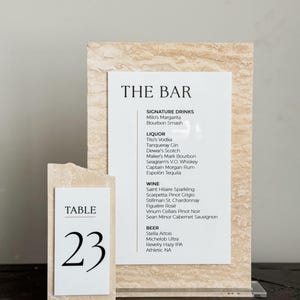 May include: A bar menu and table number 23 displayed on a textured, light-colored stone. The bar menu lists signature drinks, liquor, wine, and beer. The table number is on a separate, smaller stone.