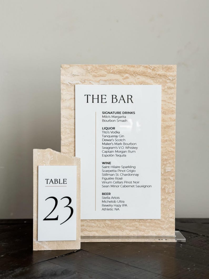 May include: A bar menu and table number 23 displayed on a textured, light-colored stone. The bar menu lists signature drinks, liquor, wine, and beer. The table number is on a separate, smaller stone.