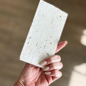 Pu&ograve; includere: Una piastrella rettangolare in pietra bianco sporco con una superficie strutturata. La piastrella &egrave; tenuta in mano, mostrando il suo aspetto poroso e macchiettato. La pietra misura circa 15 cm di lunghezza e 7,5 cm di larghezza.