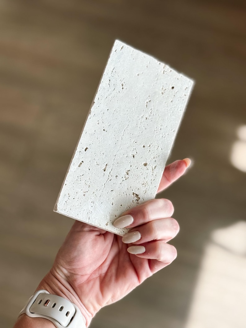 Pu&ograve; includere: Una piastrella rettangolare in pietra bianco sporco con una superficie strutturata. La piastrella &egrave; tenuta in mano, mostrando il suo aspetto poroso e macchiettato. La pietra misura circa 15 cm di lunghezza e 7,5 cm di larghezza.