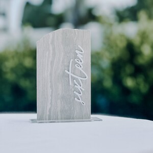 Op de afbeelding: Een rechthoekig tafelnummer met een gemarmerd grijs patroon en het woord "sixteen" in witte letters. Het tafelnummer is gemonteerd op een heldere acryl basis. De achtergrond is wazig met groen gebladerte.