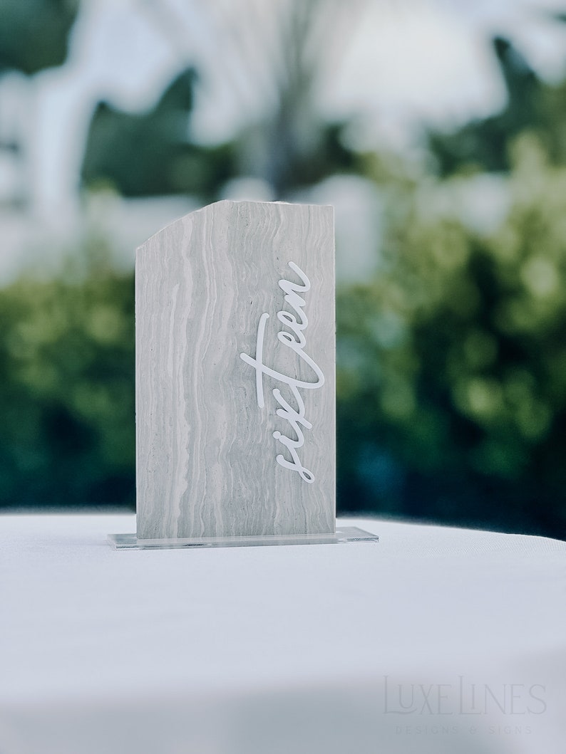 Op de afbeelding: Een rechthoekig tafelnummer met een gemarmerd grijs patroon en het woord "sixteen" in witte letters. Het tafelnummer is gemonteerd op een heldere acryl basis. De achtergrond is wazig met groen gebladerte.