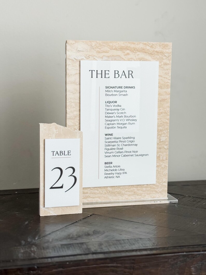 May include: A bar menu and table number 23 displayed on a wooden table. The bar menu lists signature drinks, liquor, wine, and beer. The table number is in a black font on a white background.
