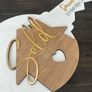 May include: A white and wood house-shaped key with the word "Sold" in gold lettering. The key has a black outline and a small heart cutout. The key is attached to a white and gold key with the text "Davin Diga 219.869.0824 | REALTOR" in black lettering.